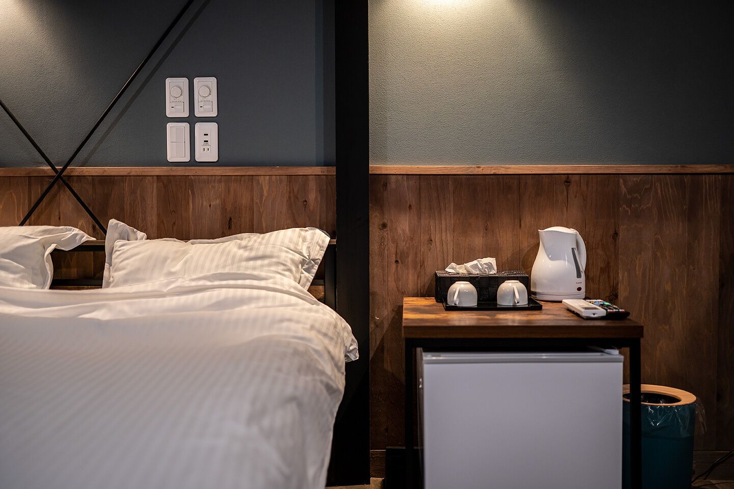 Hotellihuone, jossa on sänky, yöpöytä, jossa on kahvinkeitin, ja roskakori.
