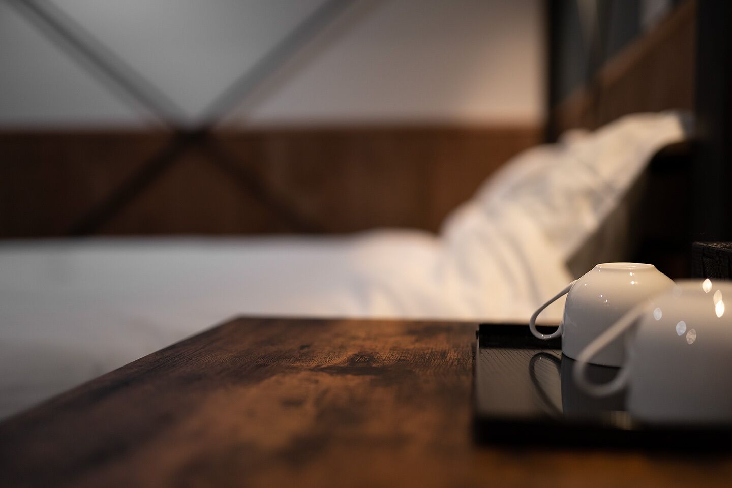 A neatly made bed with white linens and a wooden headboard. A white mug with a handle is placed on a wooden nightstand beside the bed.