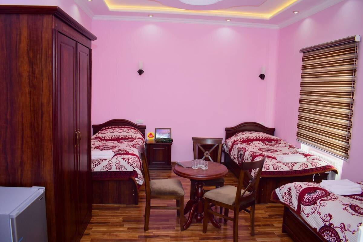 A bedroom with two beds, a wooden wardrobe, a small table with chairs, and a window with blinds.