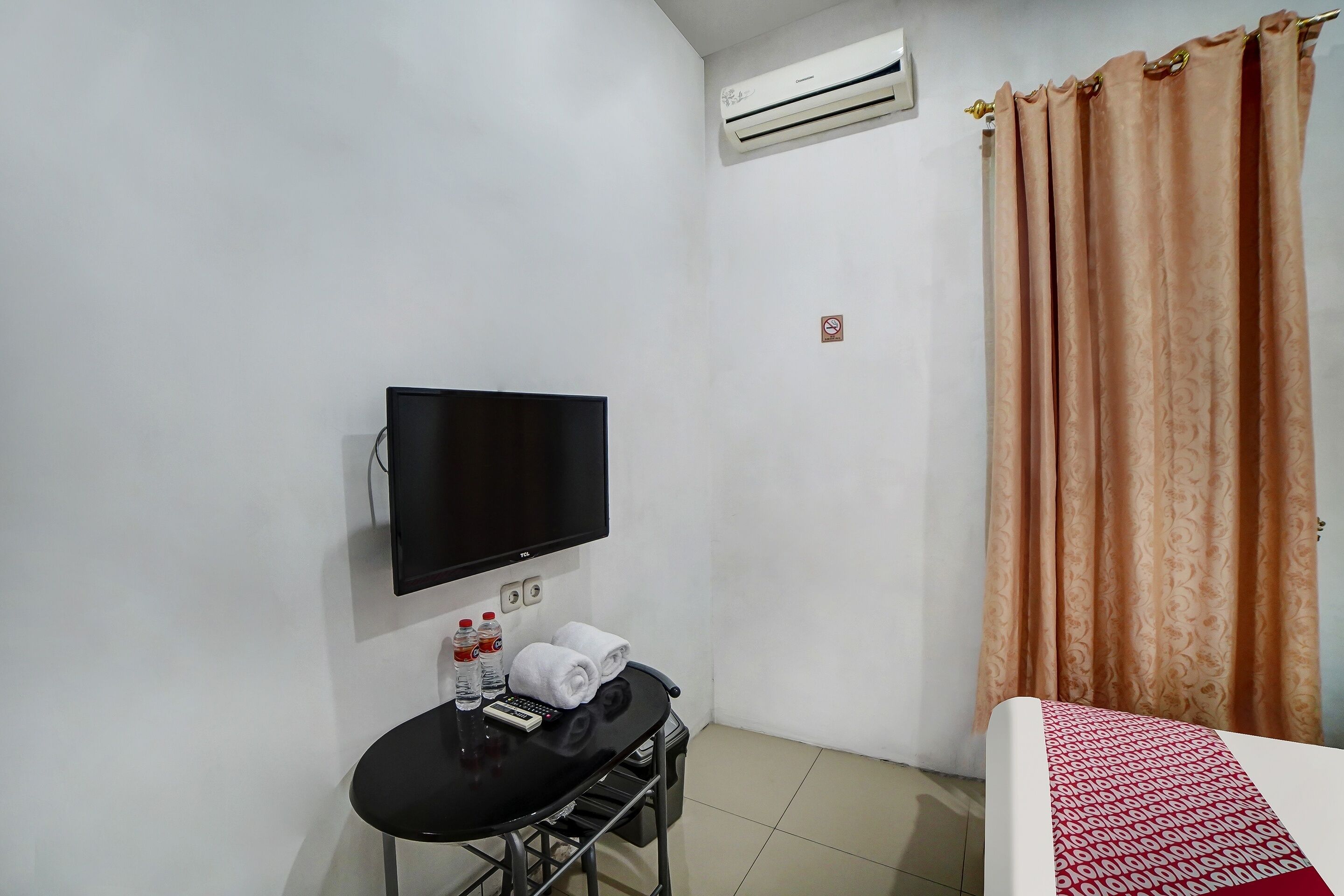 A hotel room with a bed, a TV mounted on the wall, a small table with a remote control and bottled water, and a curtain.