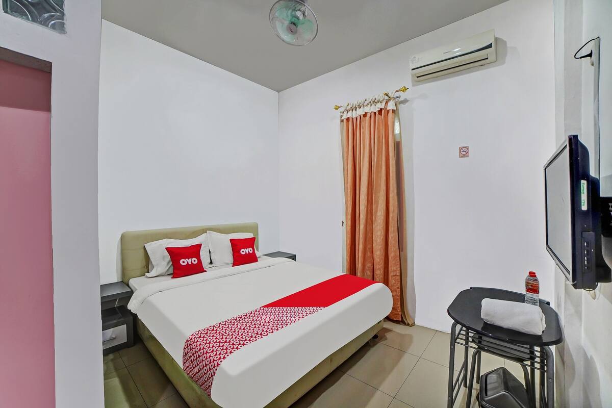 A hotel room with a bed, a TV, an air conditioner, and a small table with a towel and a bottle.