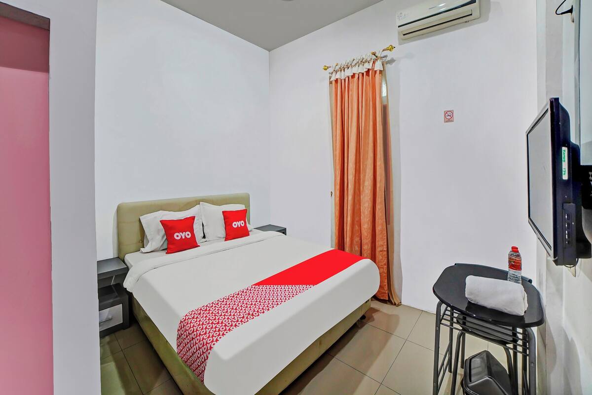 A hotel room with a bed, an air conditioner, a TV, a chair, and a curtain.