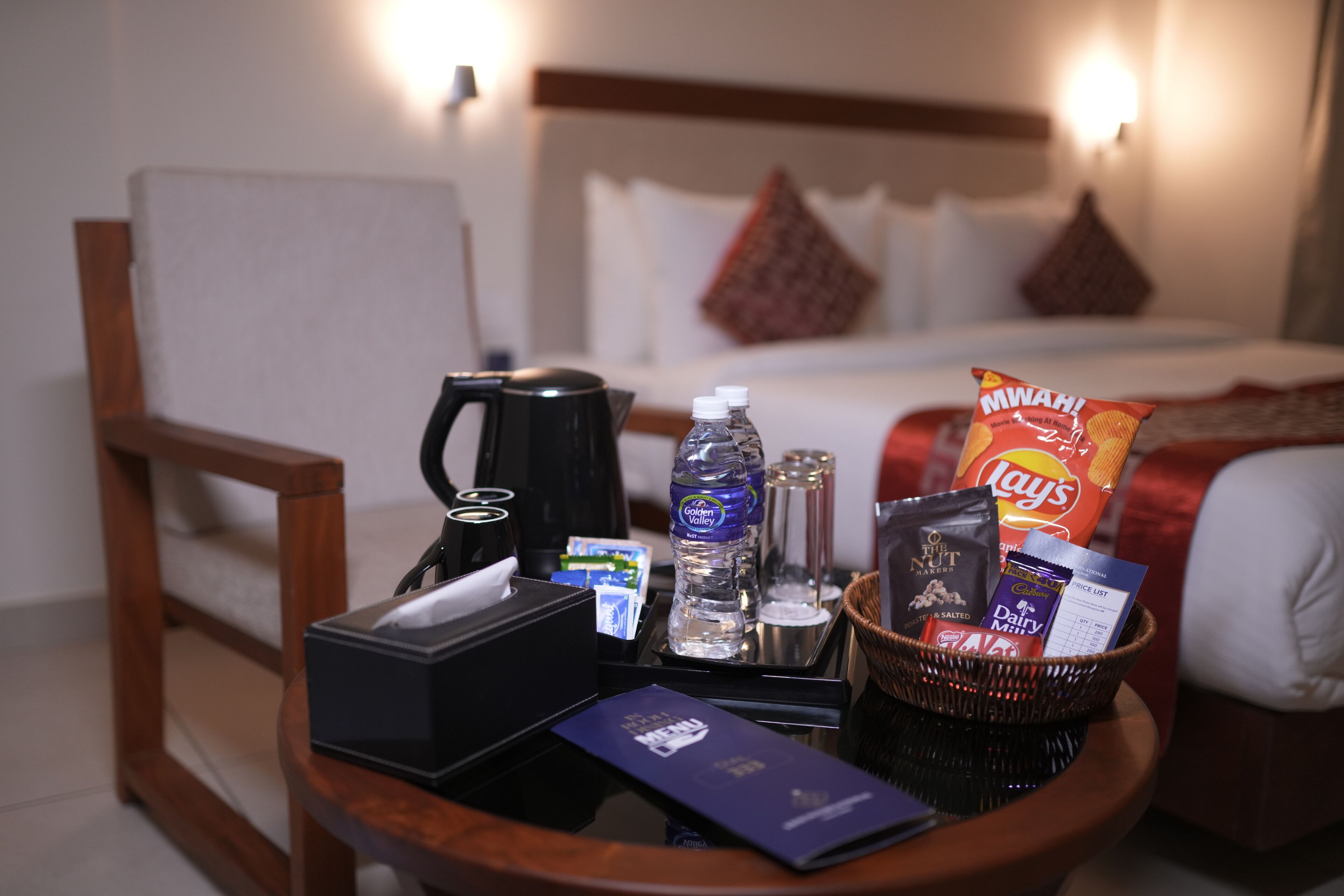 A hotel room with a bed, a bedside table with a coffee maker, water bottle, snacks, and a basket with toiletries.