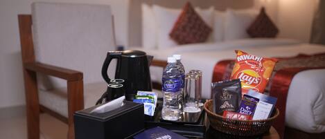 A hotel room with a bed, a bedside table with a coffee maker, water bottle, snacks, and a basket with toiletries.
