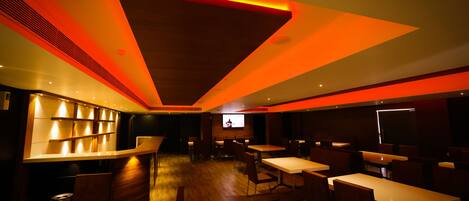 Un restaurante moderno con suelo de madera, mesas y sillas blancas. Se observa una zona de bar con estantes y una pantalla de televisión.