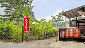 A sign for OYO hotel with a red and white design, a fenced area with banana trees, and a building with a metal roof. - OYO 91334 Permata Papua Guest House (Pameungpeuk)