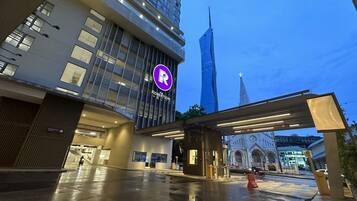 A modern hotel with a prominent 'R' logo, a covered entrance, and a view of a tall skyscraper and a church in the background.