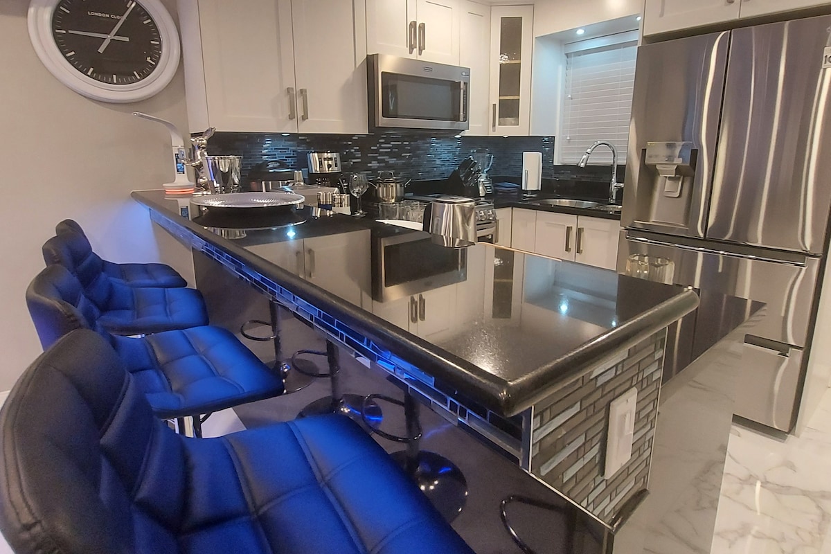 A modern kitchen with a central island, bar stools, stainless steel appliances, and a wall-mounted clock.