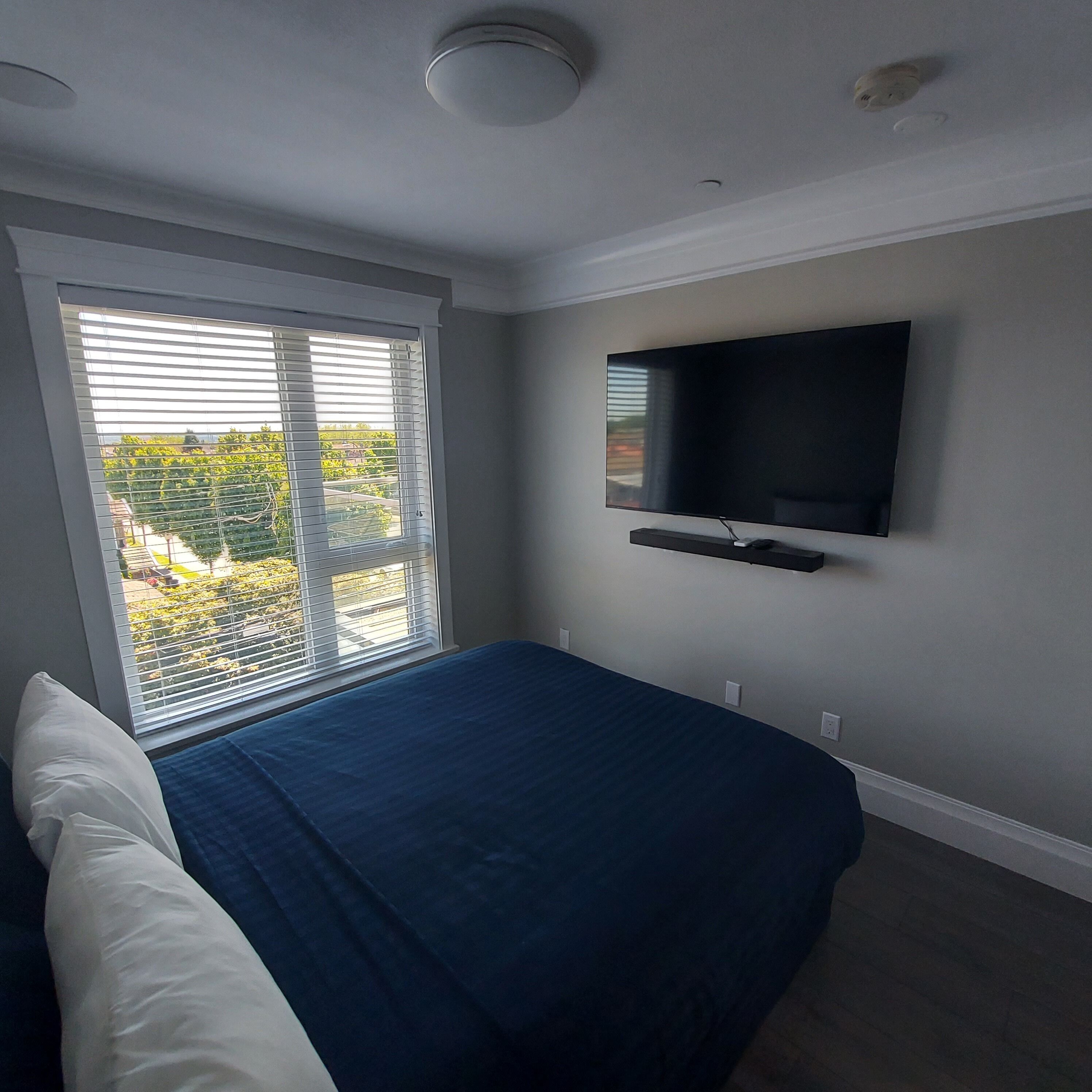 A bedroom with a bed, a TV mounted on the wall, and a window with blinds.