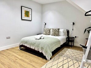 A bedroom with a bed, a bedside table, a lamp, a wall socket, and a framed picture.