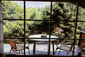 A view through a window with a table and two chairs on a wooden deck, overlooking a forested area. - Ortho Hideaway with Garden (Ortho)