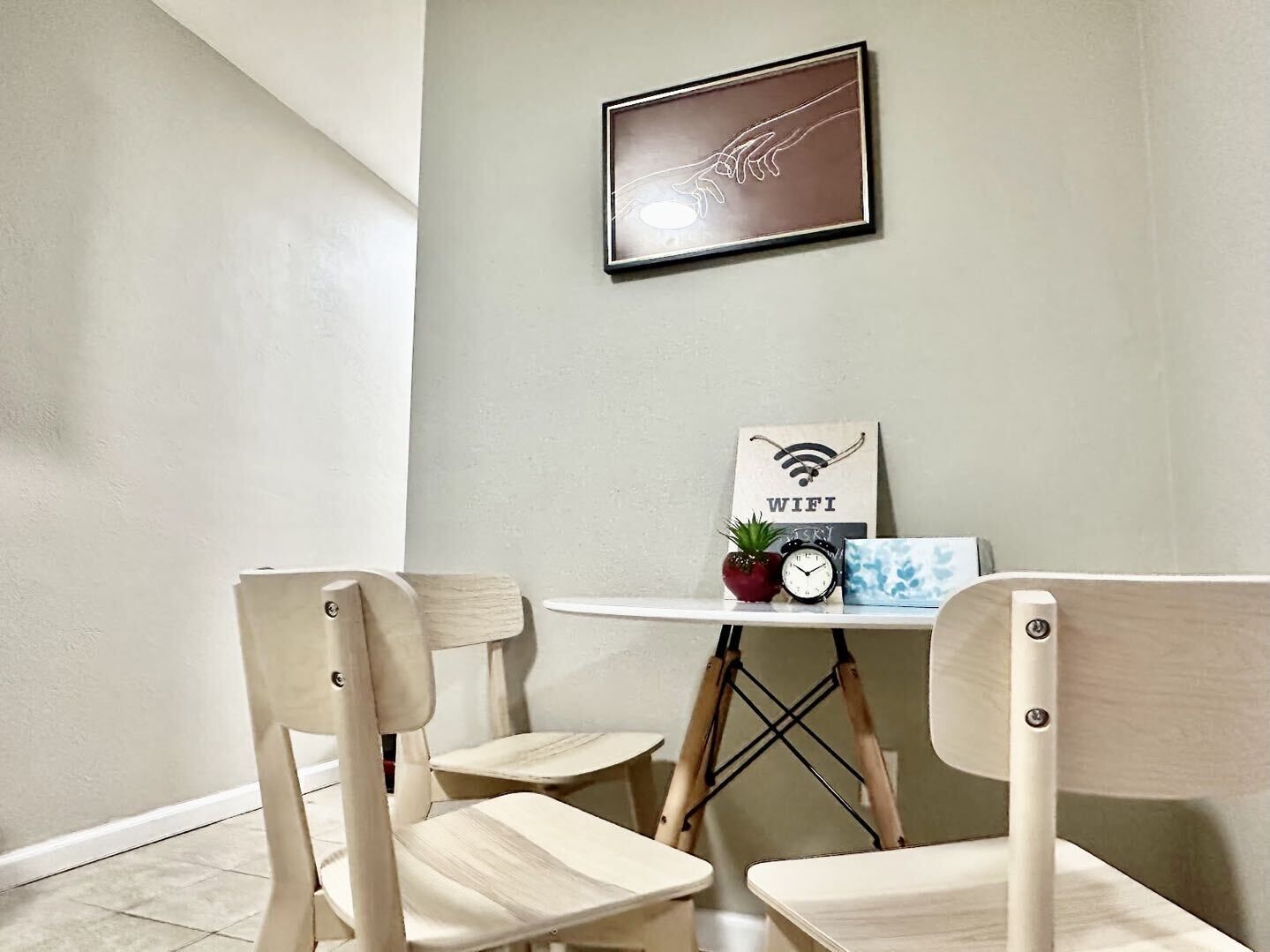 Un petit coin repas avec deux chaises en bois, une table blanche, une œuvre d’art encadrée, une petite plante en pot, une horloge et un panneau indiquant « WIFI ».