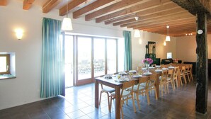 A dining area with a long wooden table set for a meal, surrounded by chairs, with a view of the outdoors through large windows. - Holiday Home in Romefort near Lac de Vassiviere (Saint Léonard de Noblat)