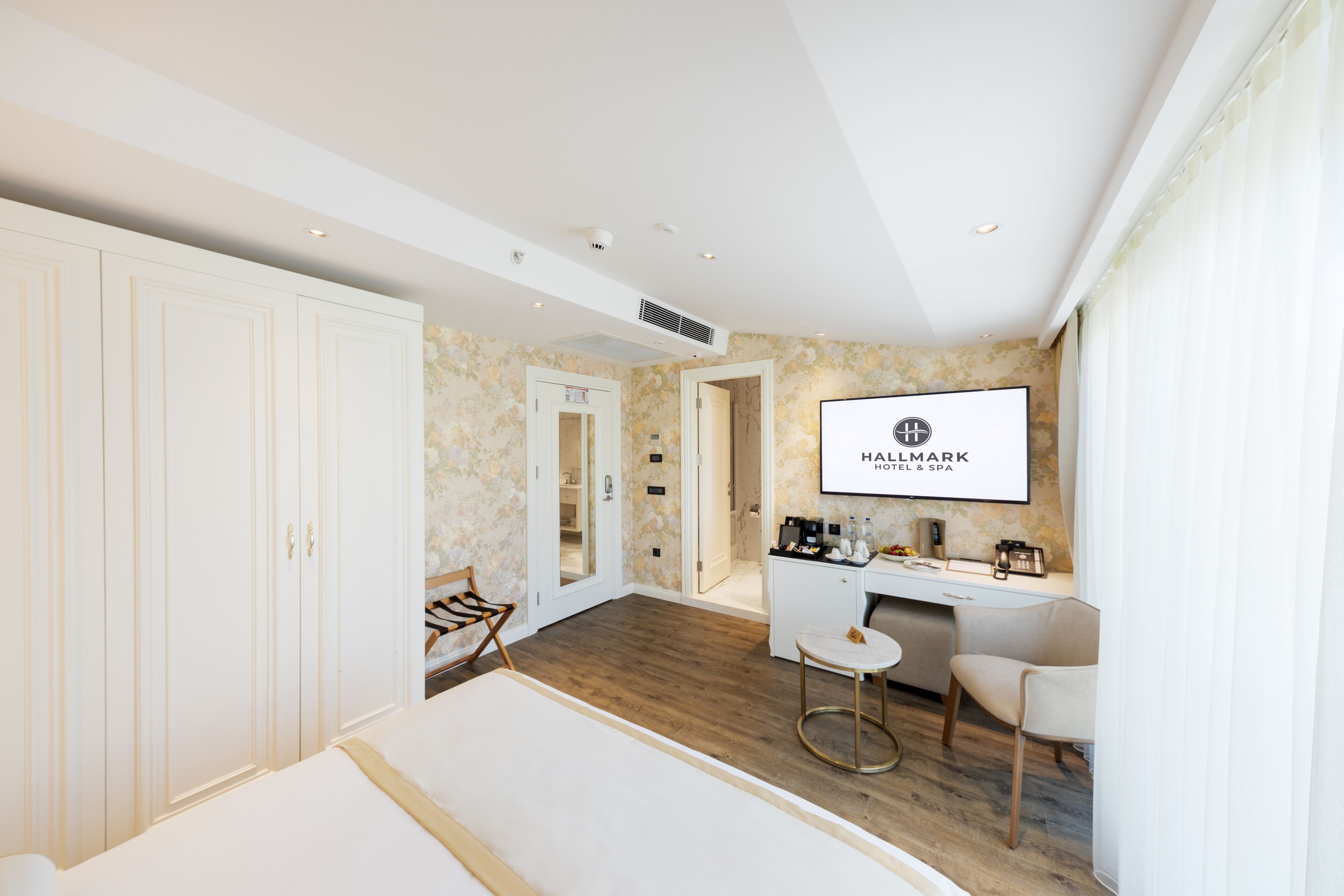 Une chambre d’hôtel avec un grand lit, un bureau, une chaise et un mini-bar. Il y a un miroir au mur et une porte qui donne sur une autre pièce.