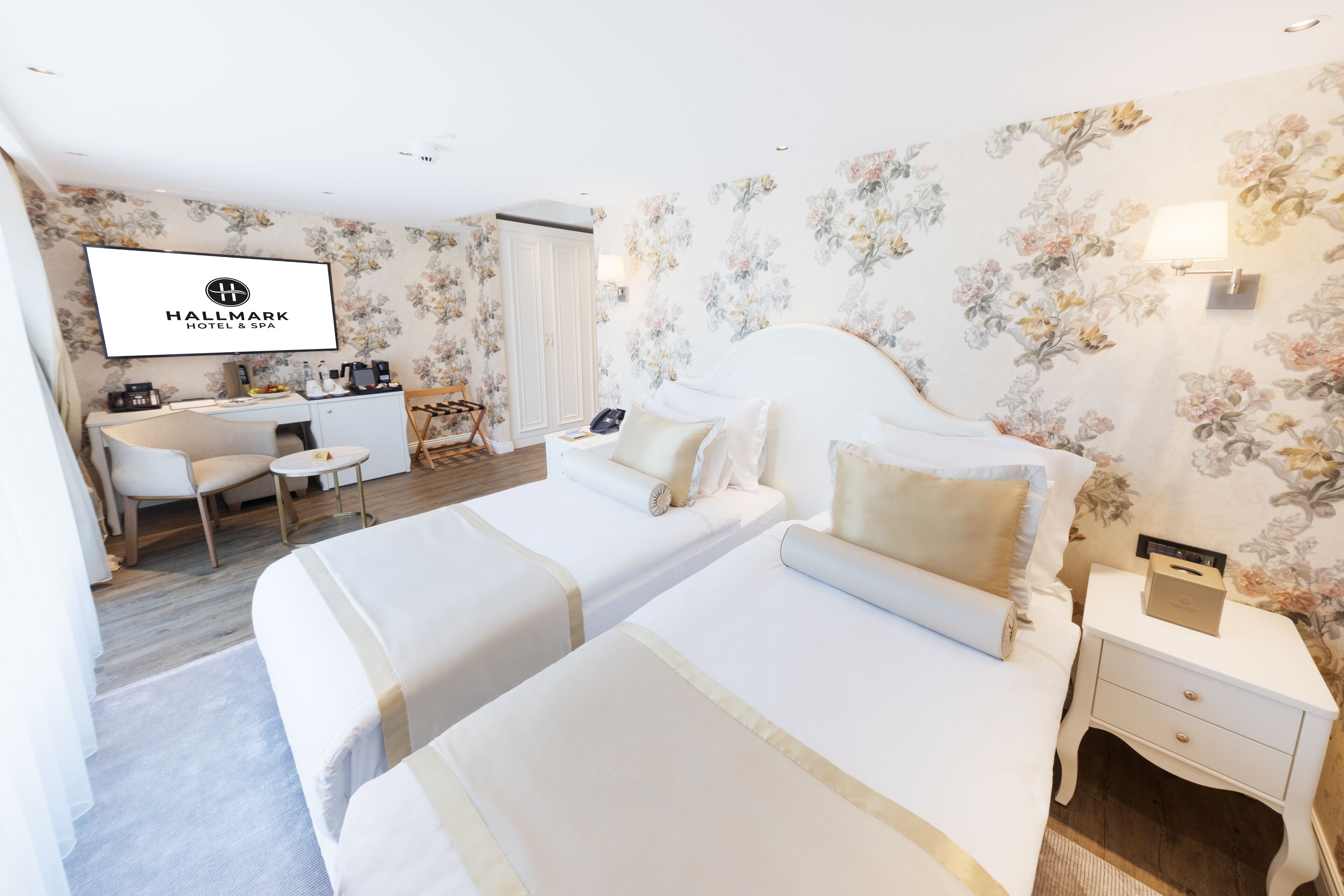 A hotel room with two beds, a desk, a chair, a TV, and a floral wallpaper.