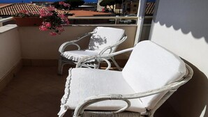 A balcony with two white wicker chairs, a glass table, and a potted plant with pink flowers.