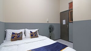 A neatly made bed with white bedding and blue and yellow patterned pillows, a standing fan, and a door with a sign on it. - SPOT ON 91761 Ramah Tamah Homestay Syariah (Pekanbaru)