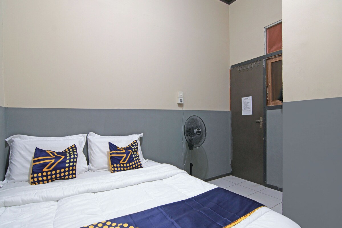 A bed with white bedding and two pillows with blue and yellow patterns, a standing fan, and a door with a window.