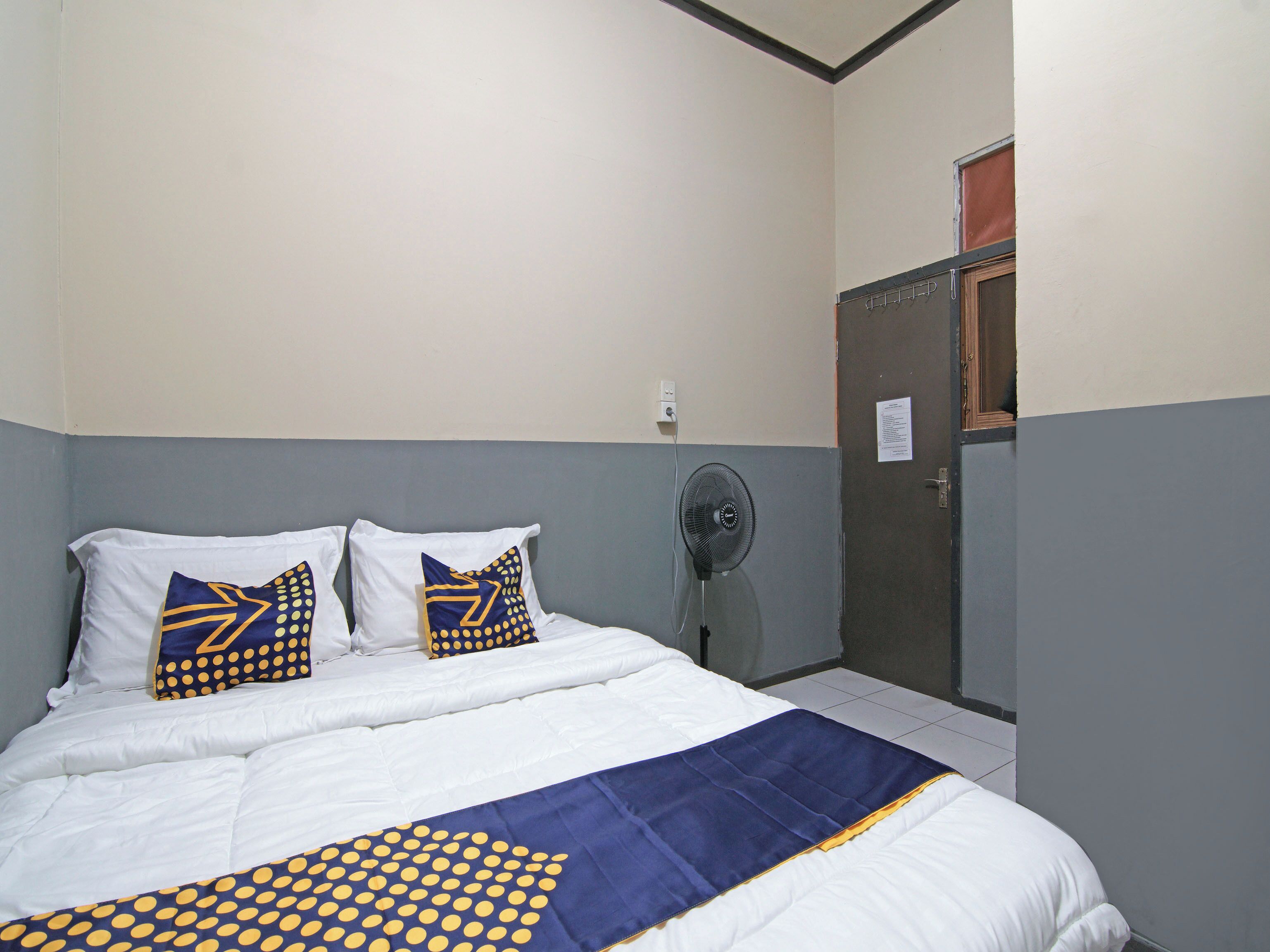 Een opgemaakt bed met witte lakens en kussens met een blauw en geel patroon, een ventilator en een deur met een bordje erop.