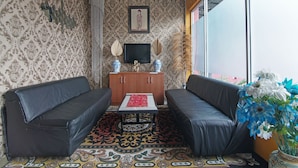 A room with patterned wallpaper, a wooden cabinet, a television, a sofa, a table, and a large window with a view. - SPOT ON 91761 Ramah Tamah Homestay Syariah (Pekanbaru)