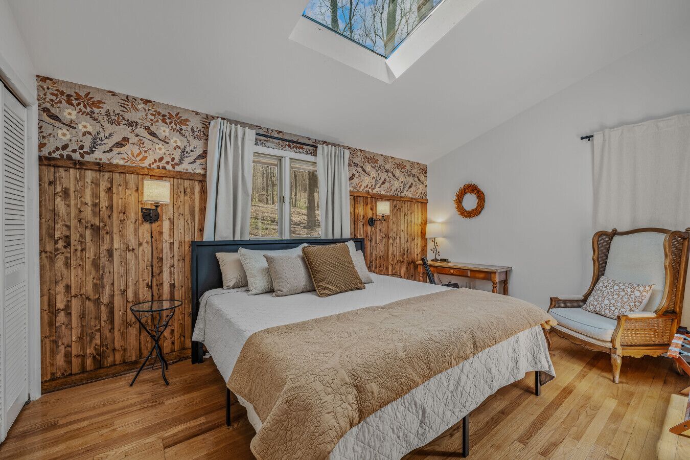 Une chambre avec un grand lit, des murs en bois, une lucarne, une chaise et une petite table.