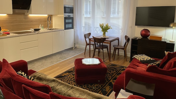 A modern living room with red sofas, a dining table with chairs, a kitchen with white cabinets, and a flat-screen TV mounted on the wall.