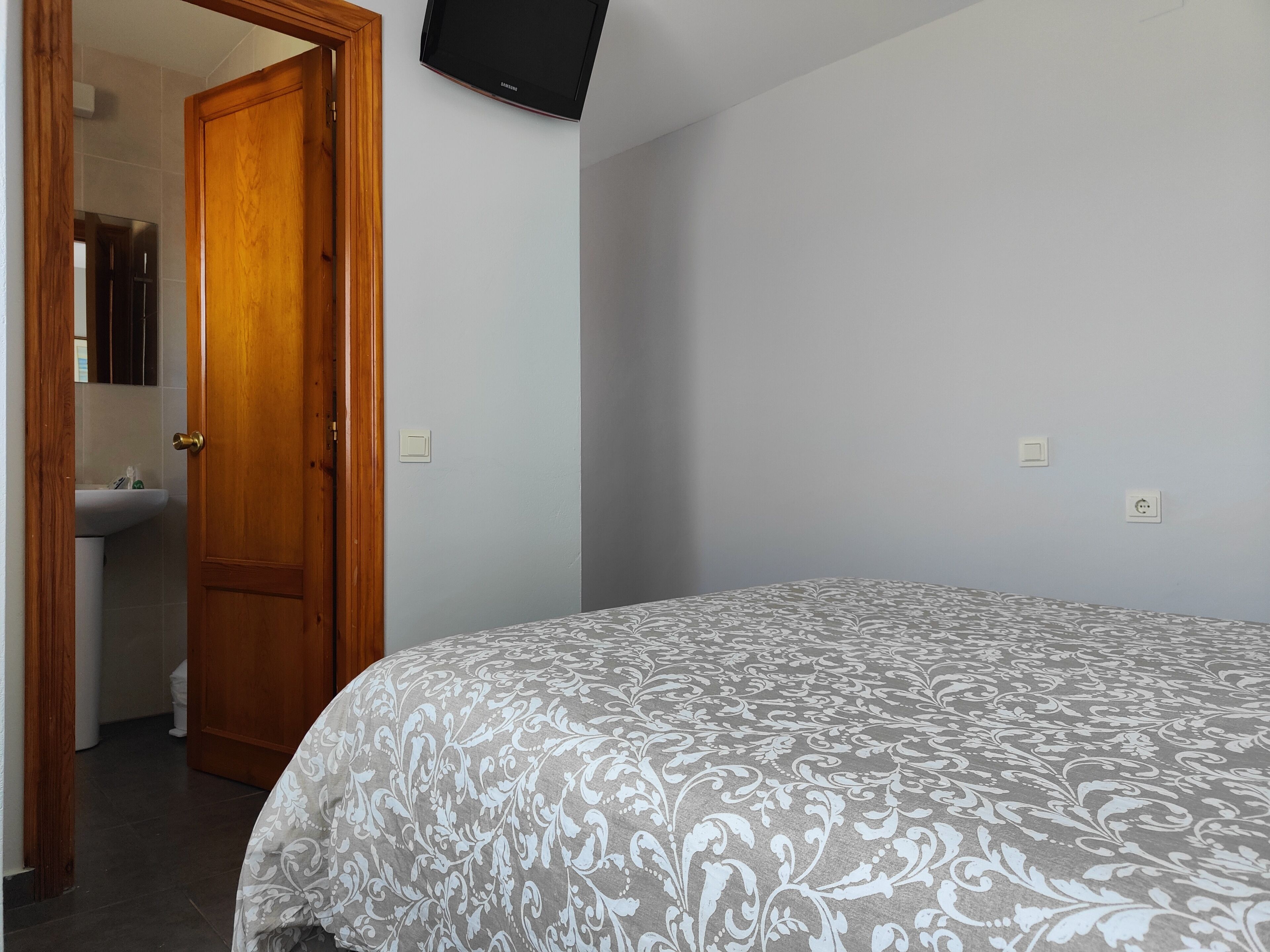 Un dormitorio con una cama, una puerta de madera, un televisor montado en la pared y un baño visible a través de una puerta abierta.
