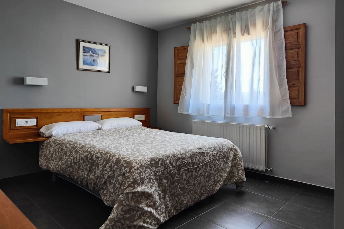 Une chambre à coucher avec une tête de lit en bois, un lit recouvert d’une literie à motifs, une table de chevet, une lampe fixée au mur, un tableau encadré et une fenêtre ornée de rideaux légers.