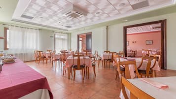 Un grand espace de salle à manger avec plusieurs tables et chaises, un coin cuisine doté d’un comptoir et de tabourets, et une porte de sortie bien visible.
