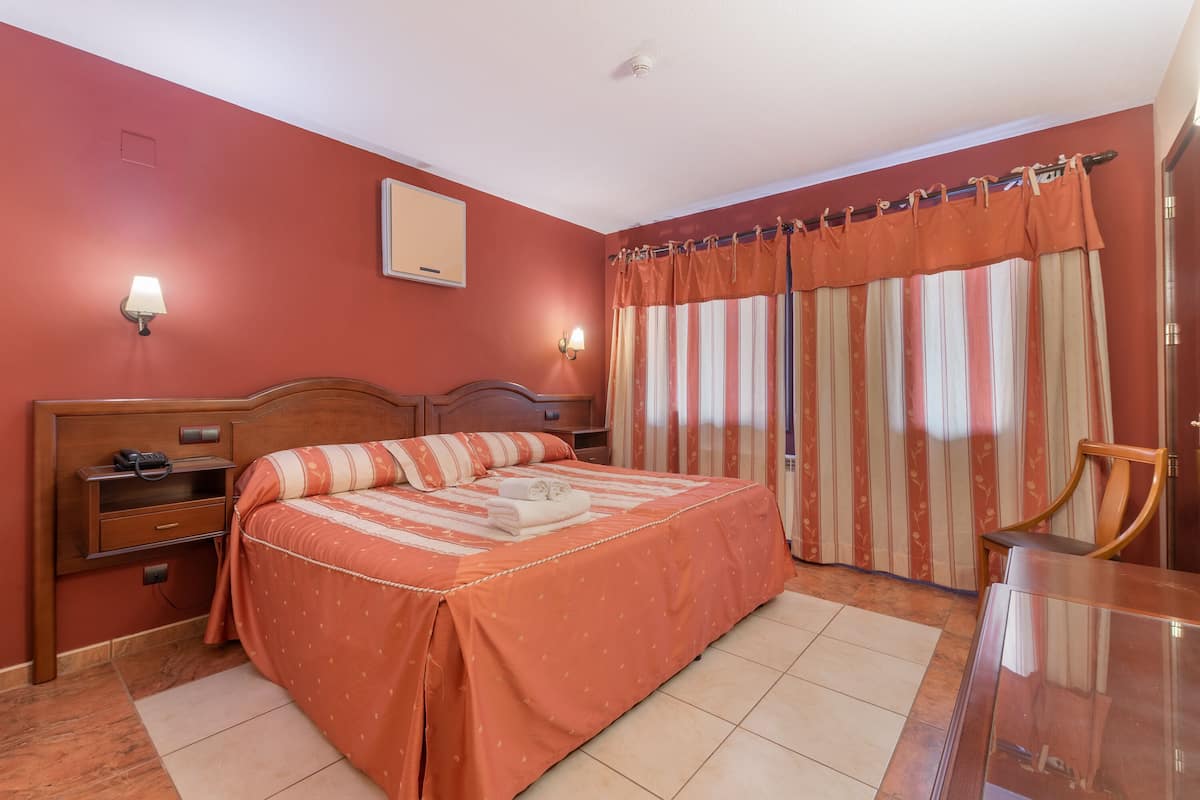 Une chambre à coucher avec un lit, une table de chevet, une lampe, une chaise et une fenêtre avec des rideaux.