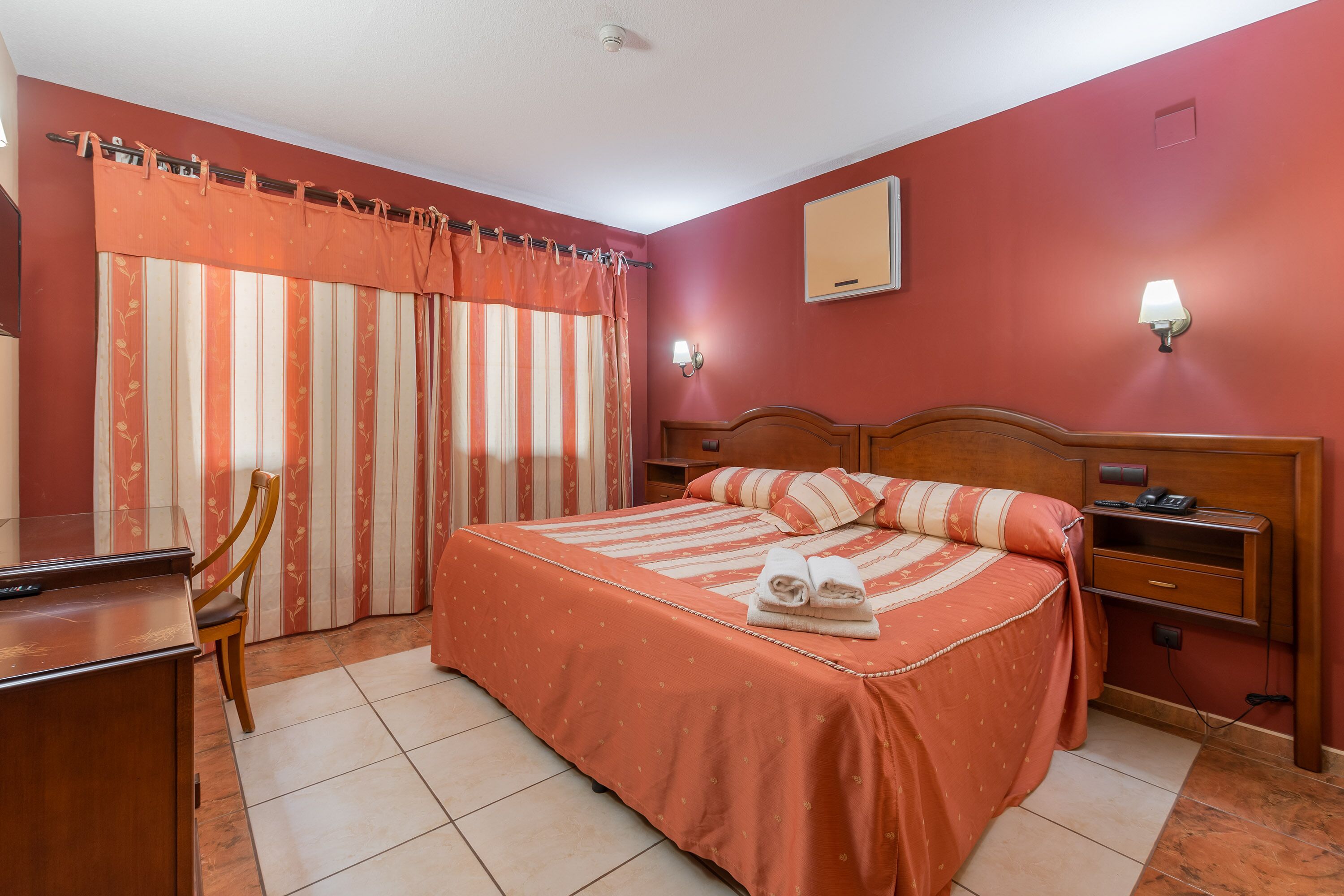 A hotel room with a bed, a desk, a chair, a nightstand, and a lamp.