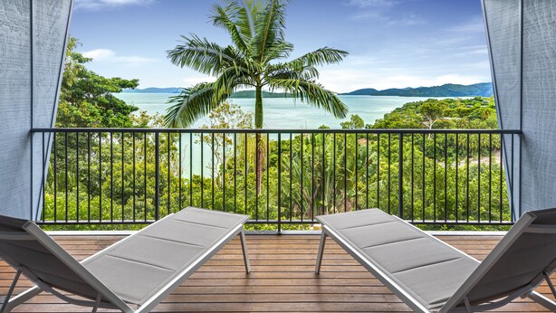 Un balcón con dos sillones de descanso, vista a un paisaje tropical con una palmera y un cuerpo de agua.