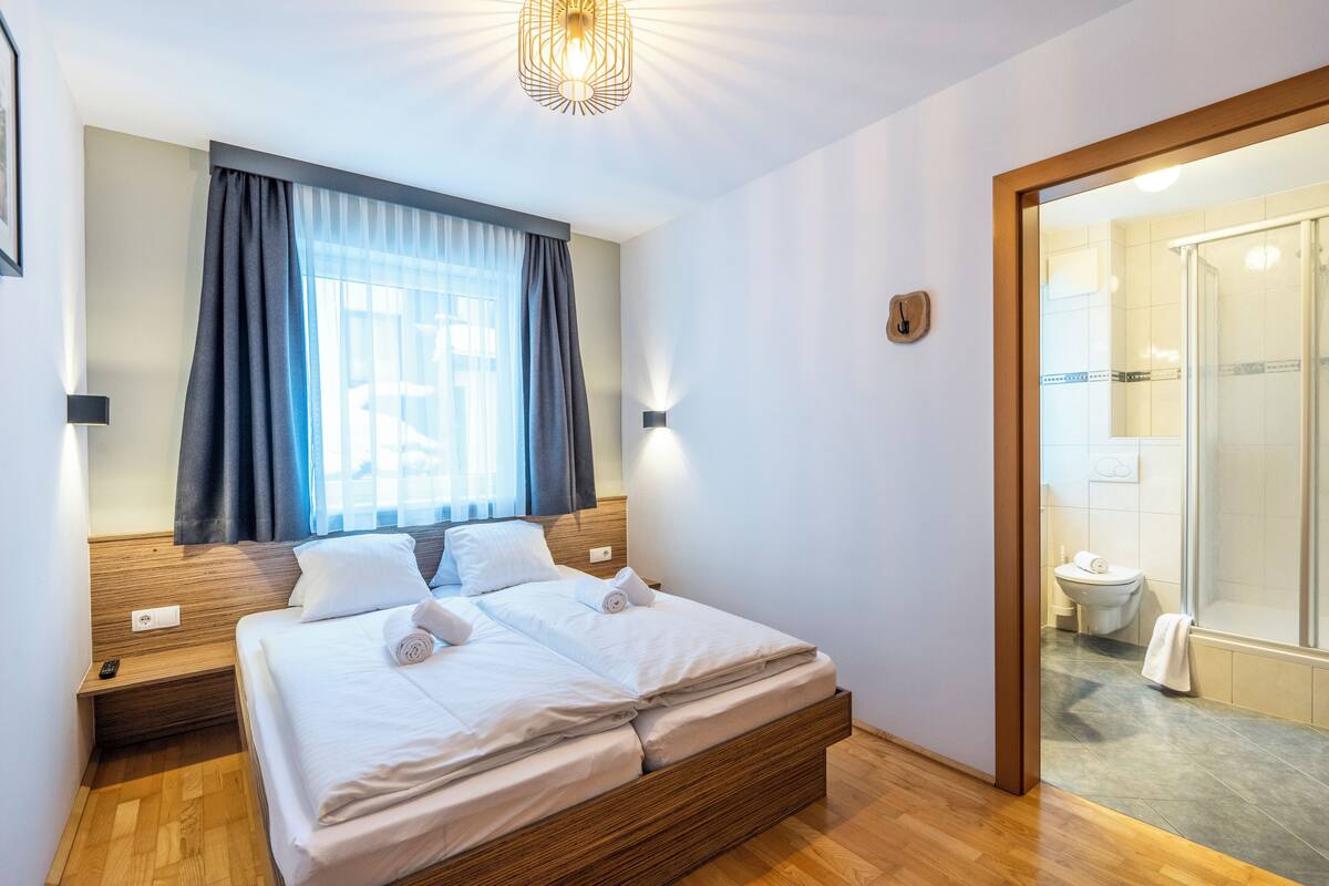 A double bed with white linens, a wooden headboard, a bedside table with a lamp, and a window with curtains.