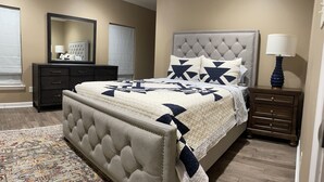 A bedroom with a tufted headboard, a large bed with a quilt, a nightstand with a lamp, a dresser, and a mirror.