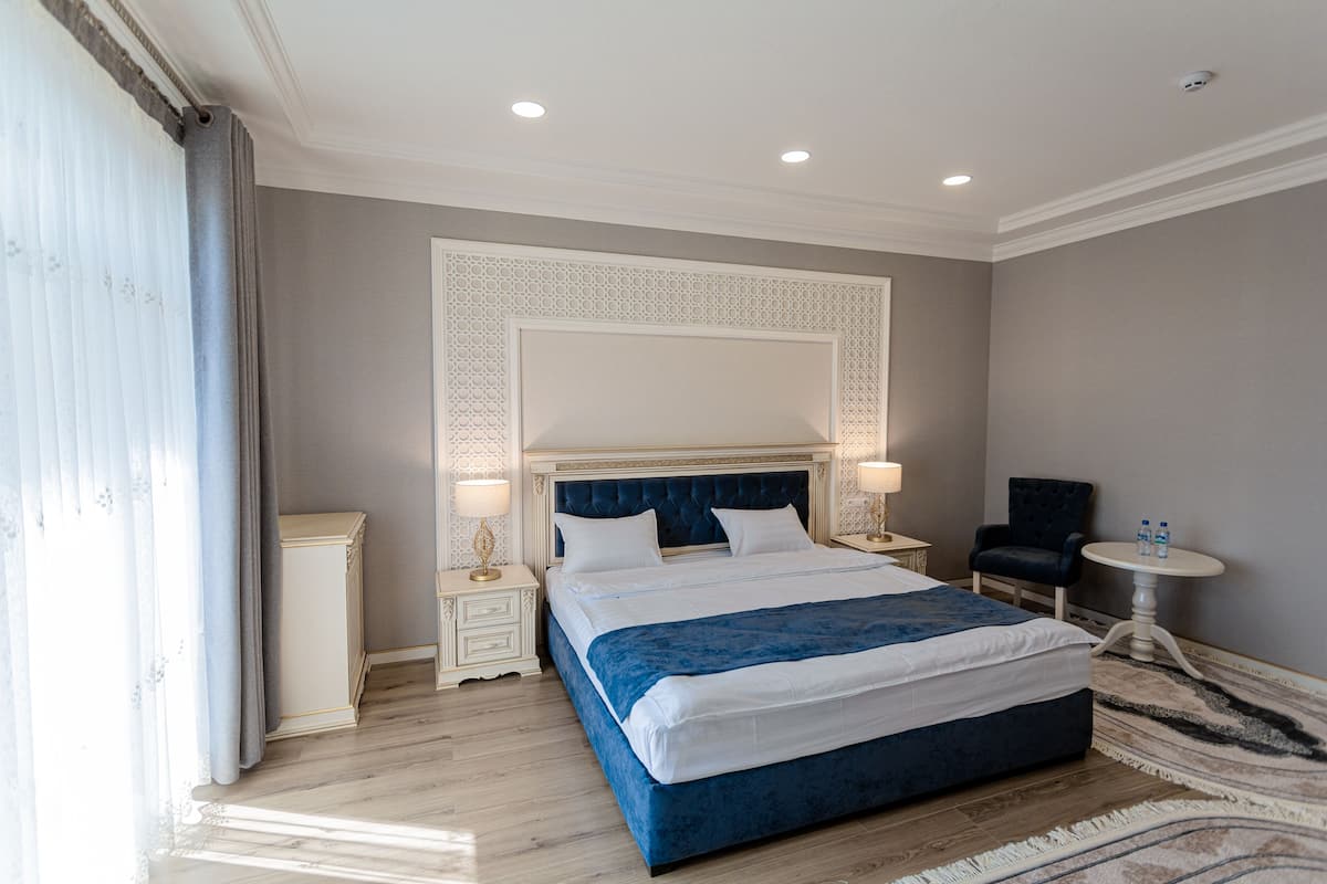 A modern hotel room with a large bed, two bedside tables with lamps, a chair, and a small round table with water bottles.