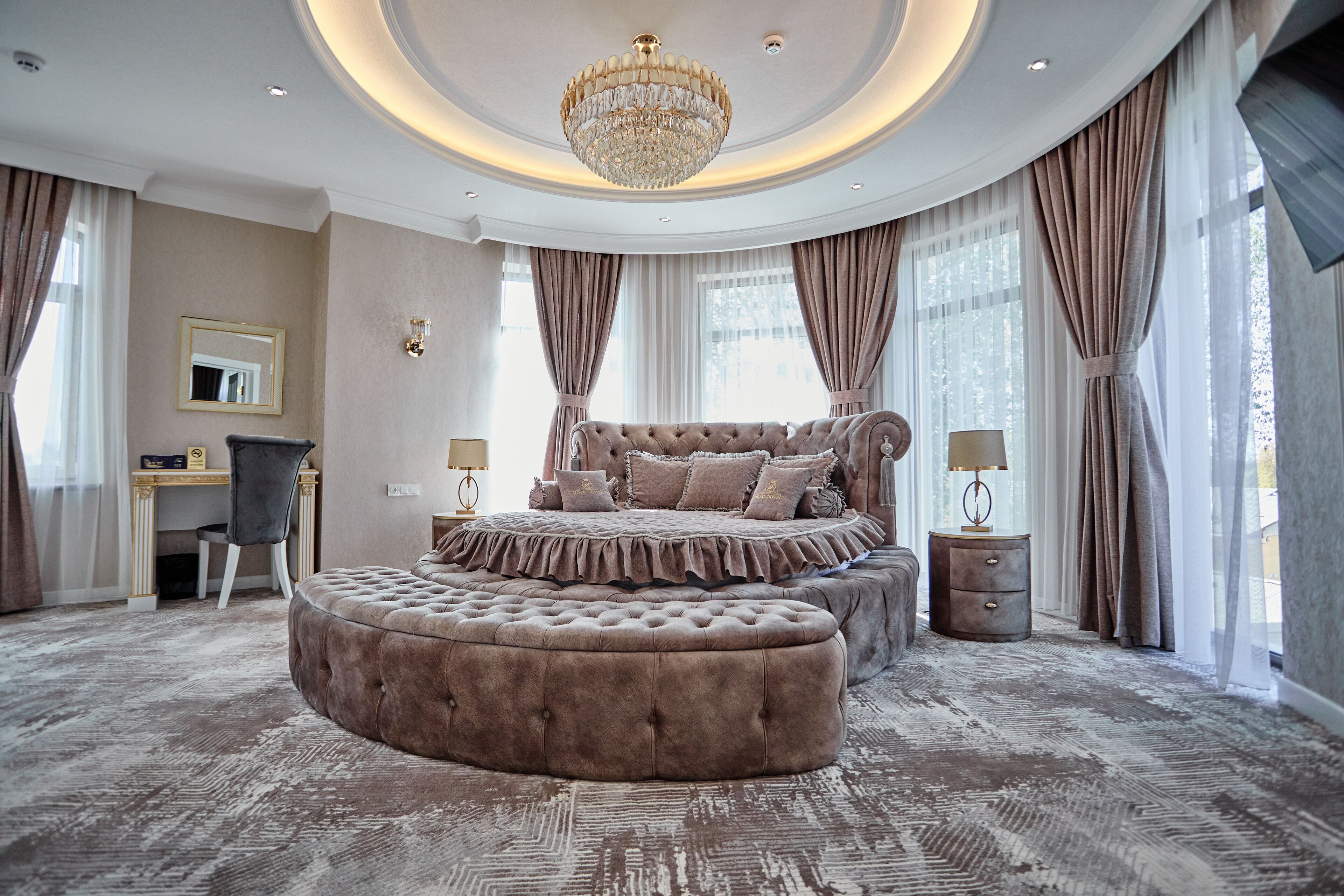 Ein luxuriöses Schlafzimmer mit einem großen, gepolsterten Bett, einem runden Hocker, einem Schminktisch mit Spiegel und einer Kristallleuchtre.
