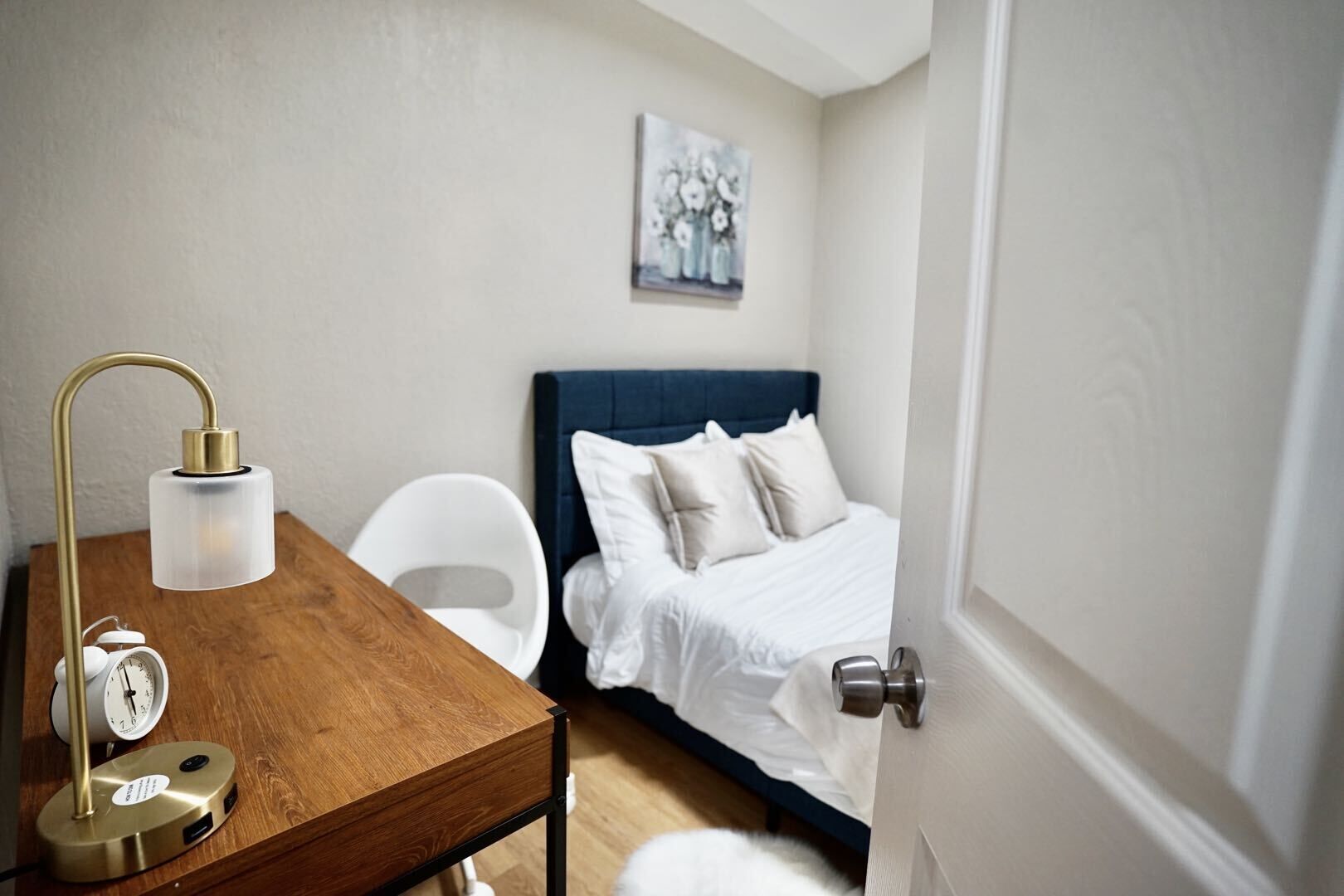 A bedroom with a wooden desk, a white chair, a bed with a blue headboard, and a painting on the wall.