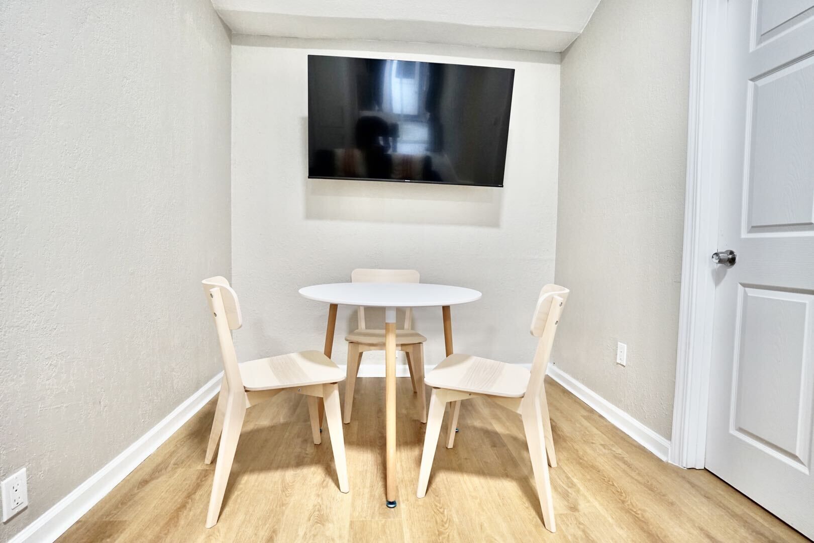 Ein kleines Zimmer mit einer weißen Tür, einer weißen Wand und einem Holzboden. In der Mitte steht ein runder Tisch mit vier Stühlen und darüber an der Wand ist ein Flachbildfernseher angebracht.