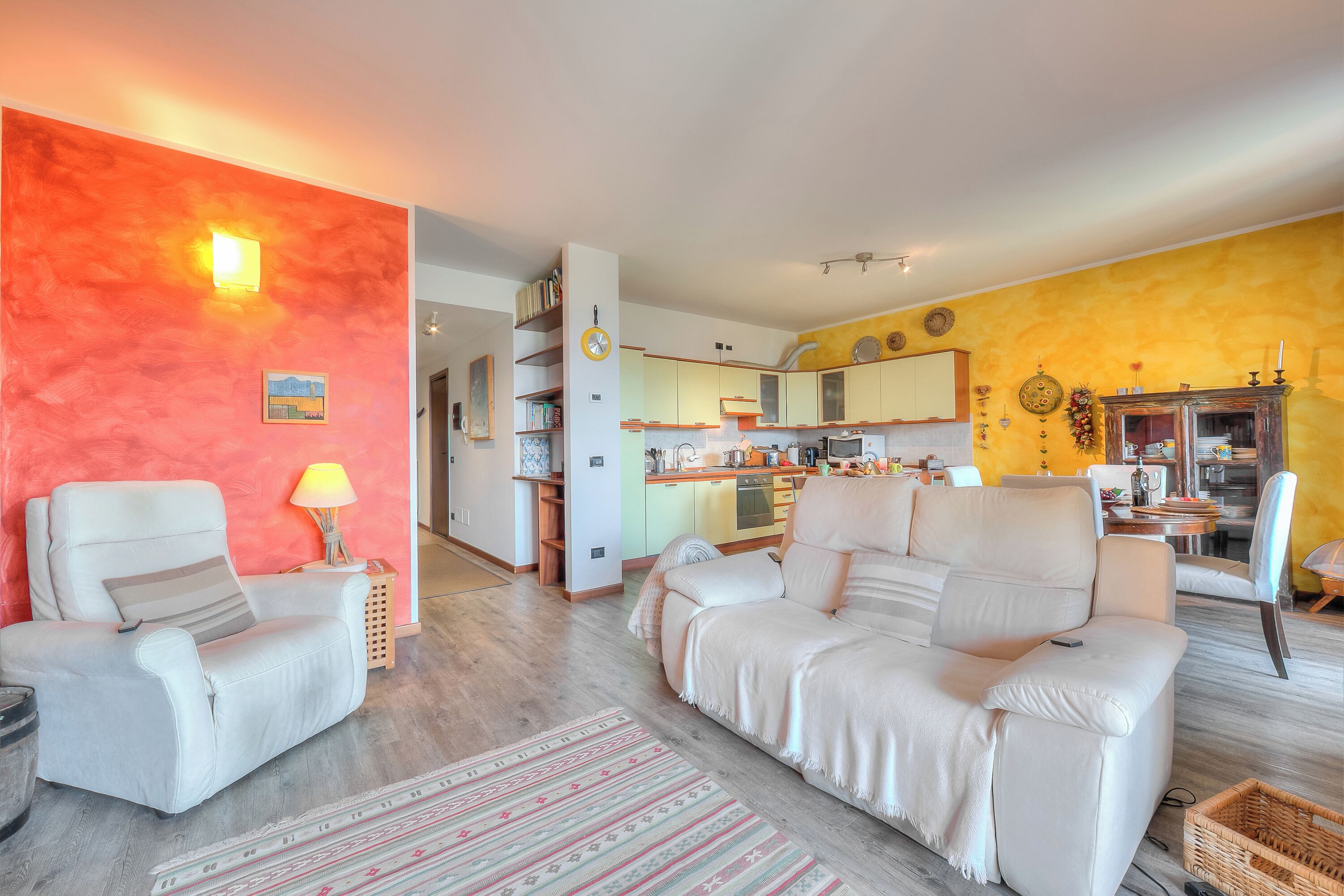 Un salon avec un canapé, des fauteuils, une lampe, un tapis et, au fond, une cuisine.