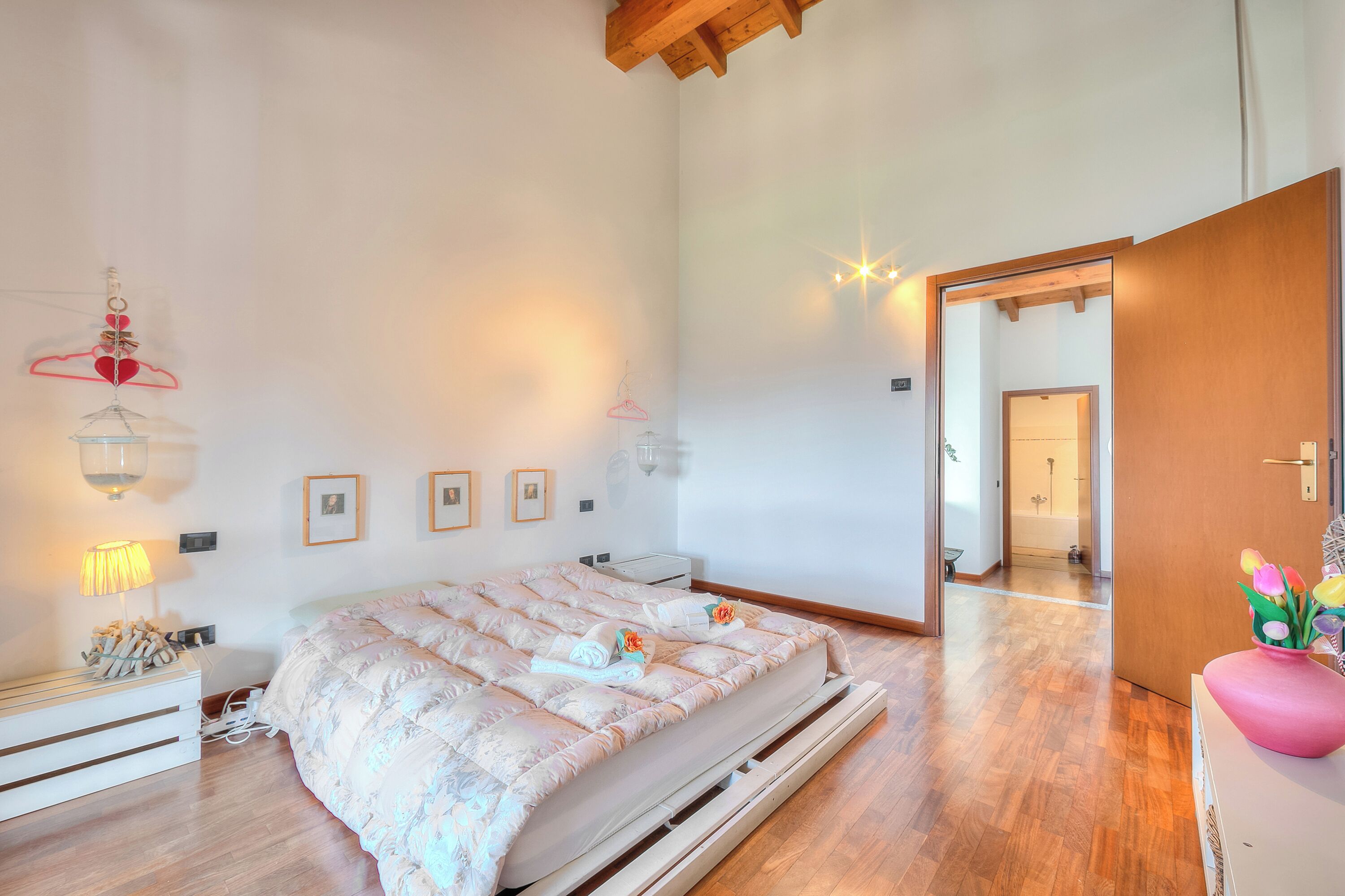 Ein Schlafzimmer mit Bett, Nachttisch, Lampe, Wandleuchten, gerahmten Bildern und Holzboden.