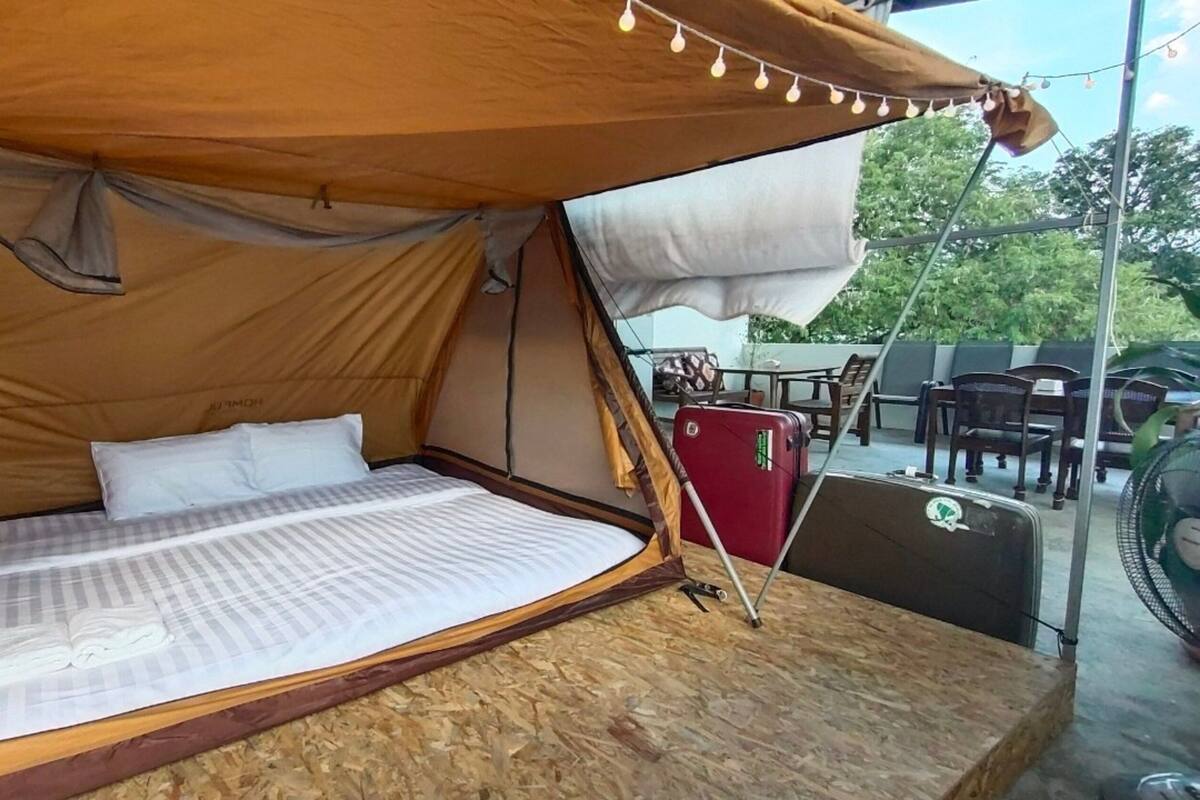 Una carpa con una cama en su interior, una maleta roja y un ventilador en el exterior.
