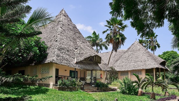 A traditional thatched-roof building with a staircase leading to a porch, surrounded by lush greenery and palm trees. - Coral Beach Resort (Diani Beach)