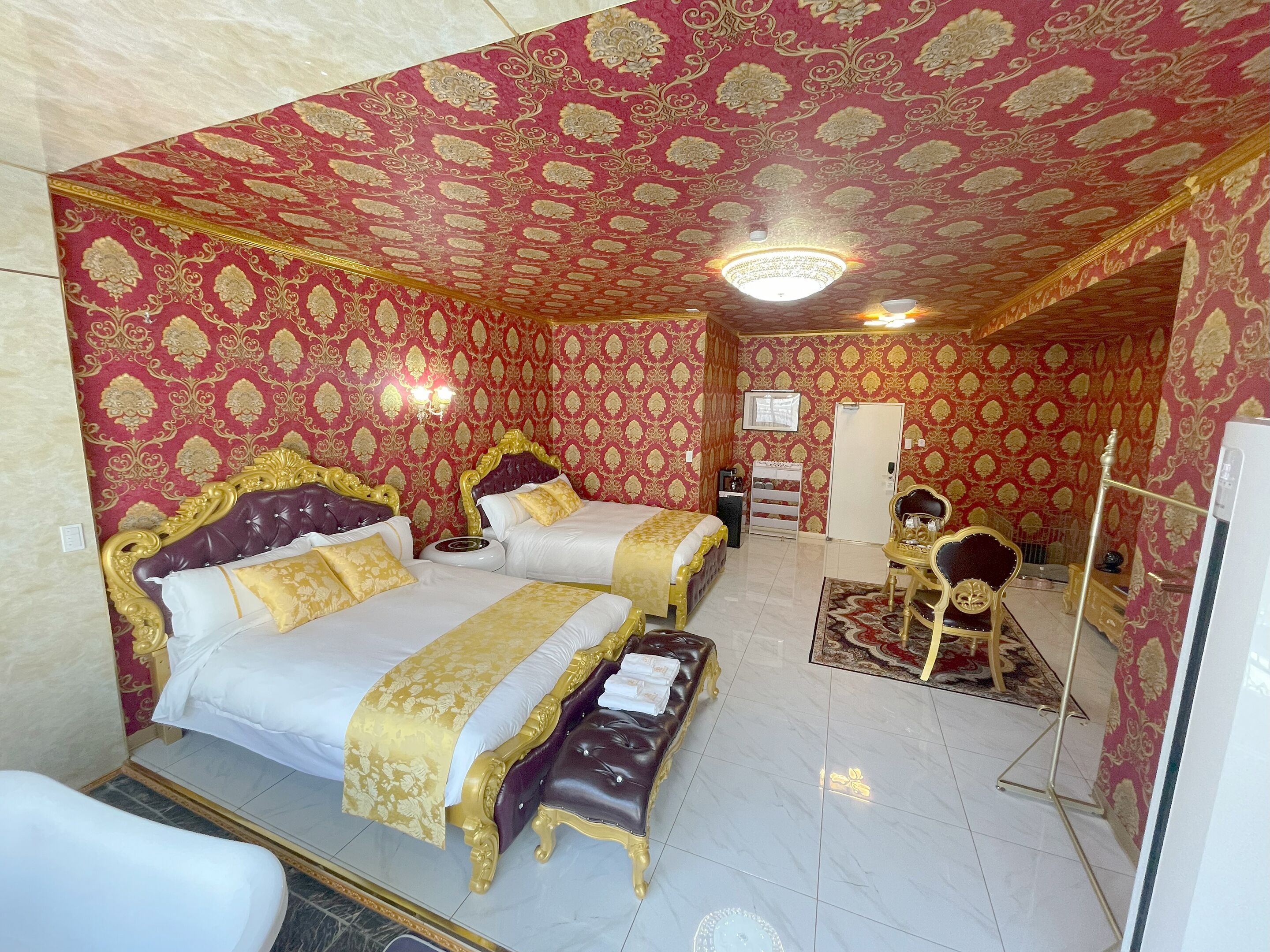 Um quarto luxuoso com papel de parede elaborado, uma cama grande, uma cama menor, uma toucador e um espelho.