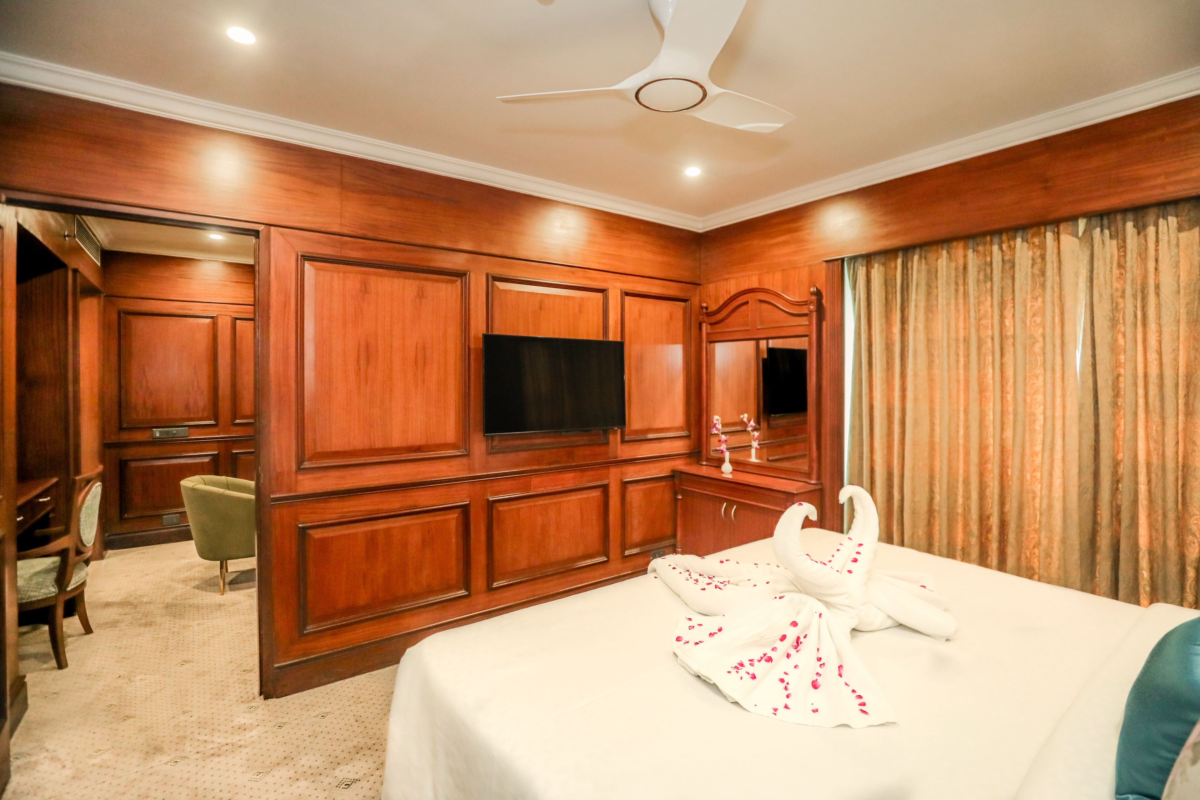 A hotel room with wooden paneling, a bed with white bedding, a swan-shaped pillow, a flat-screen TV, and a ceiling fan.