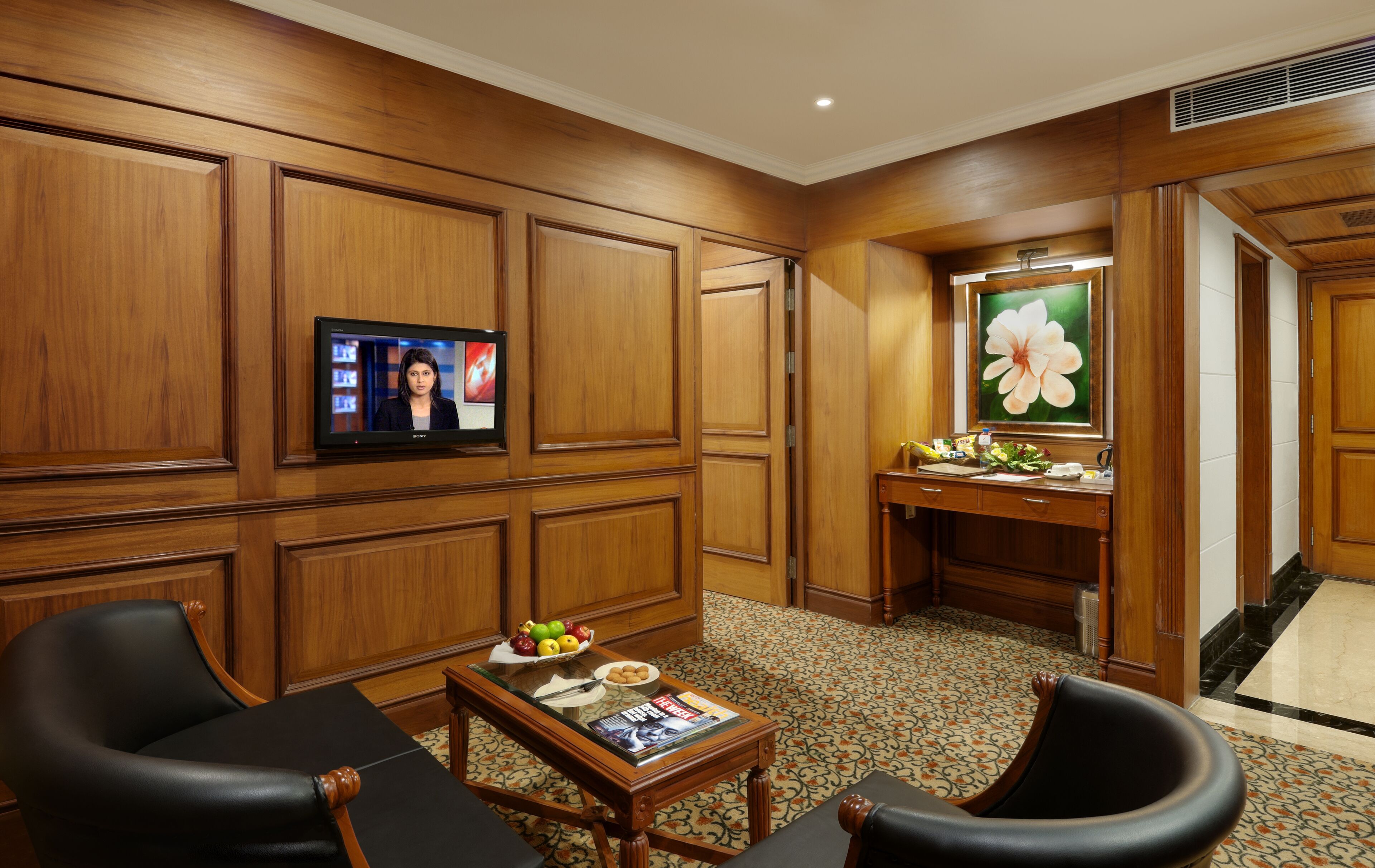 A hotel room with wooden paneling, a television, a desk with a vase of flowers, and a magazine on the table.
