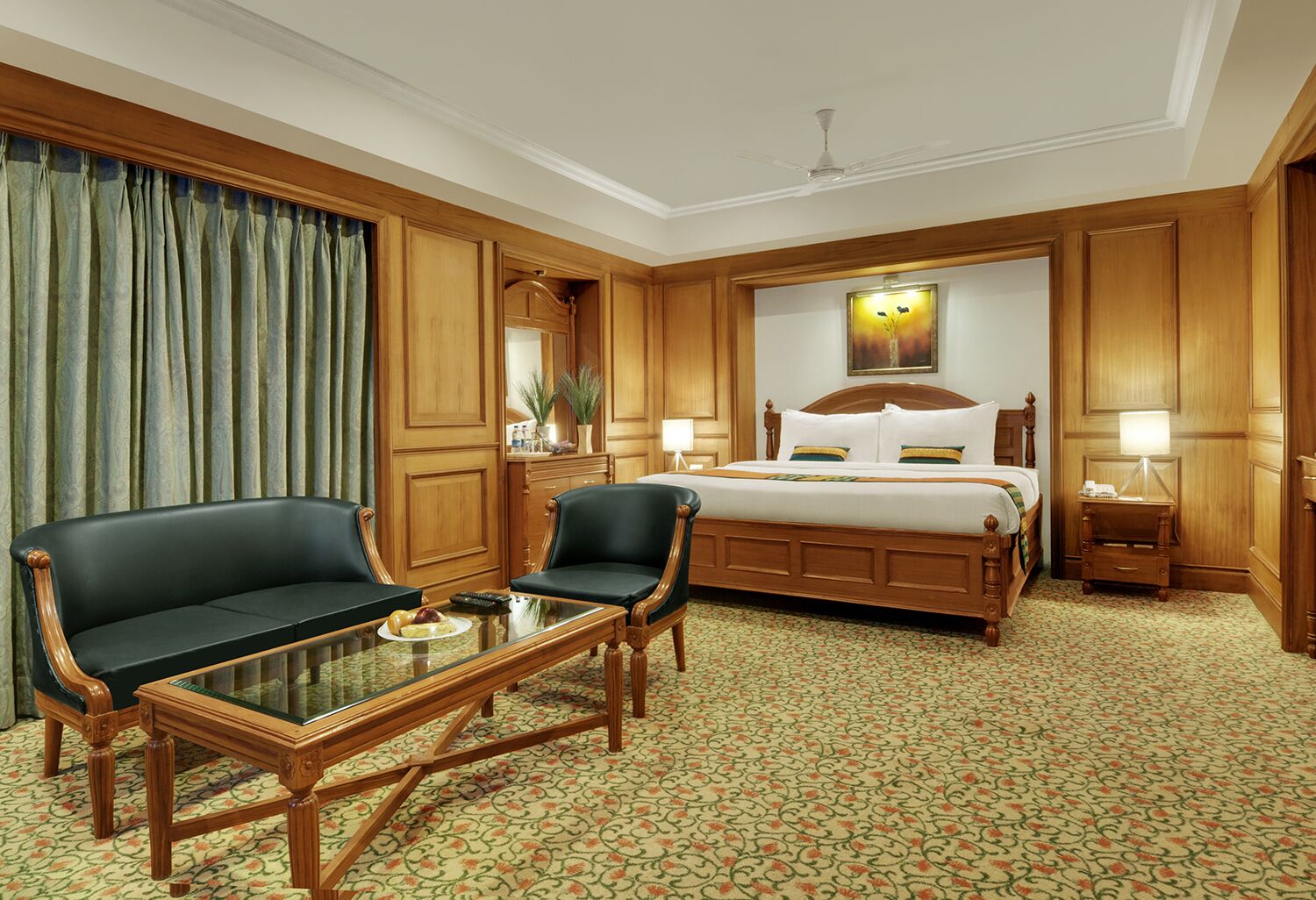 A hotel room with a large bed, two armchairs, a glass coffee table, wooden paneling, a patterned carpet, and a ceiling fan.