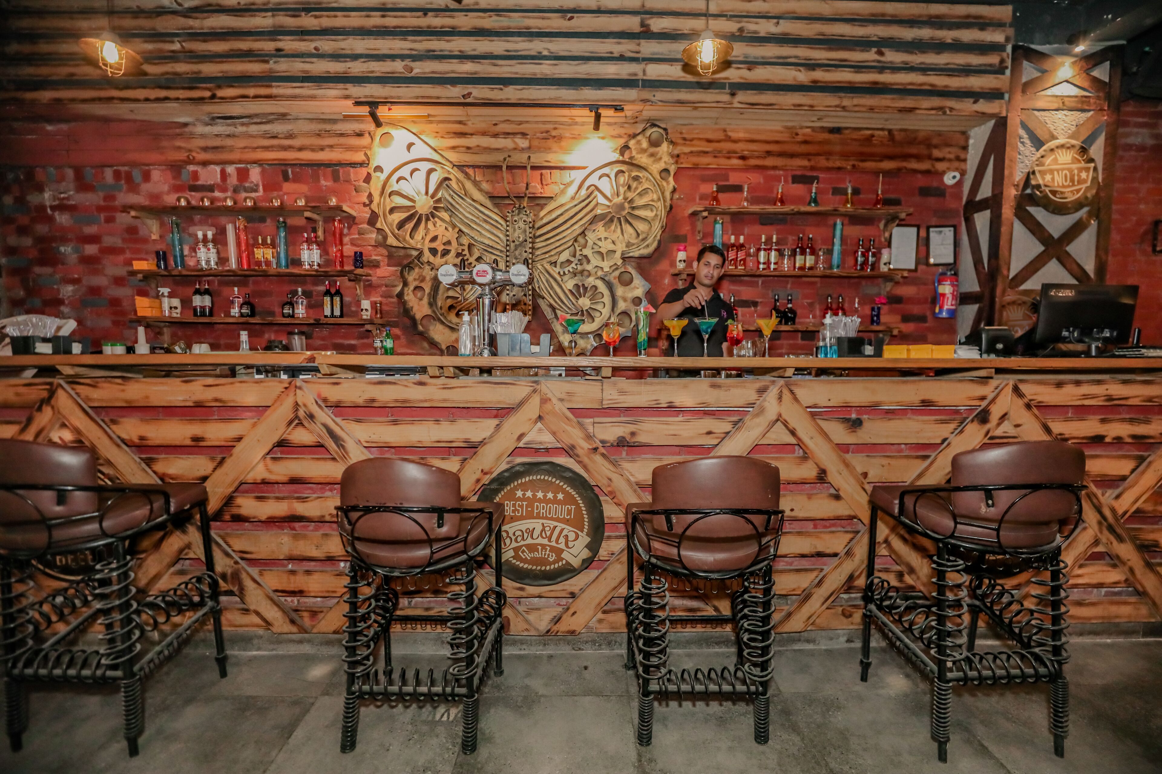 A bar with a wooden counter, shelves with bottles, and a large butterfly decoration. There are bar stools and a sign that says "Best Product Quality."