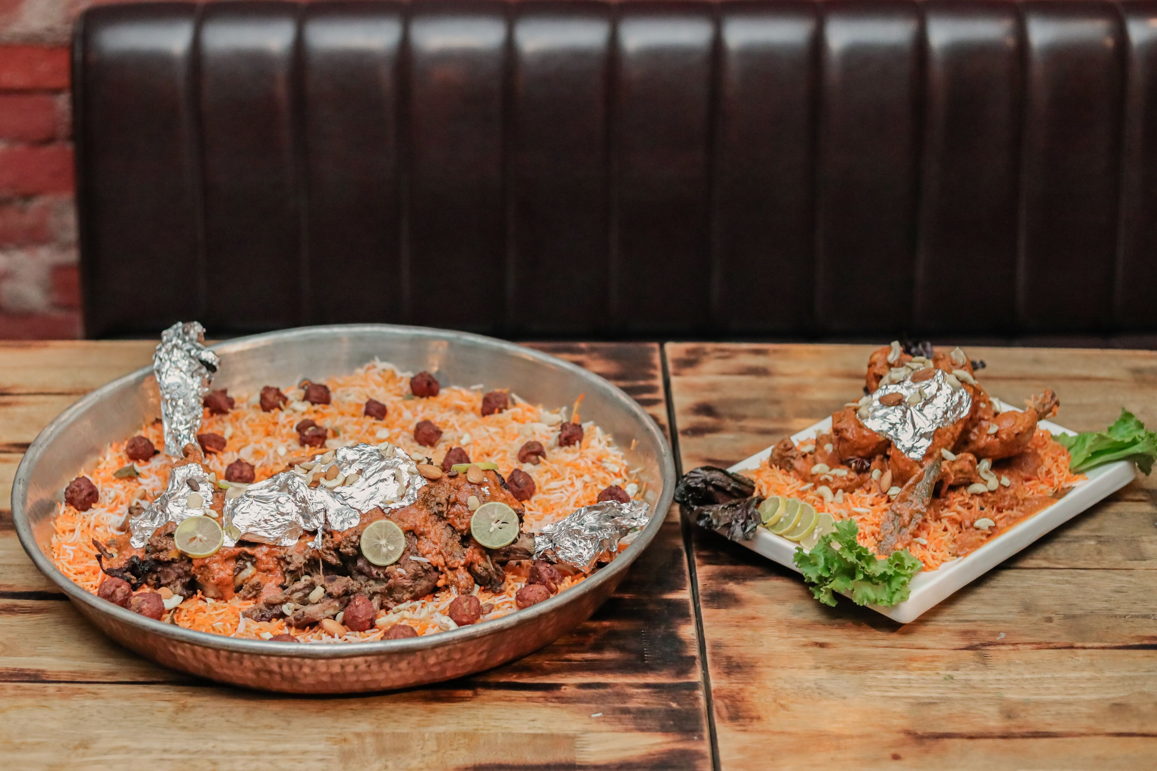 A large copper serving dish filled with rice, meat, and nuts, accompanied by a smaller plate with a piece of meat and greens.