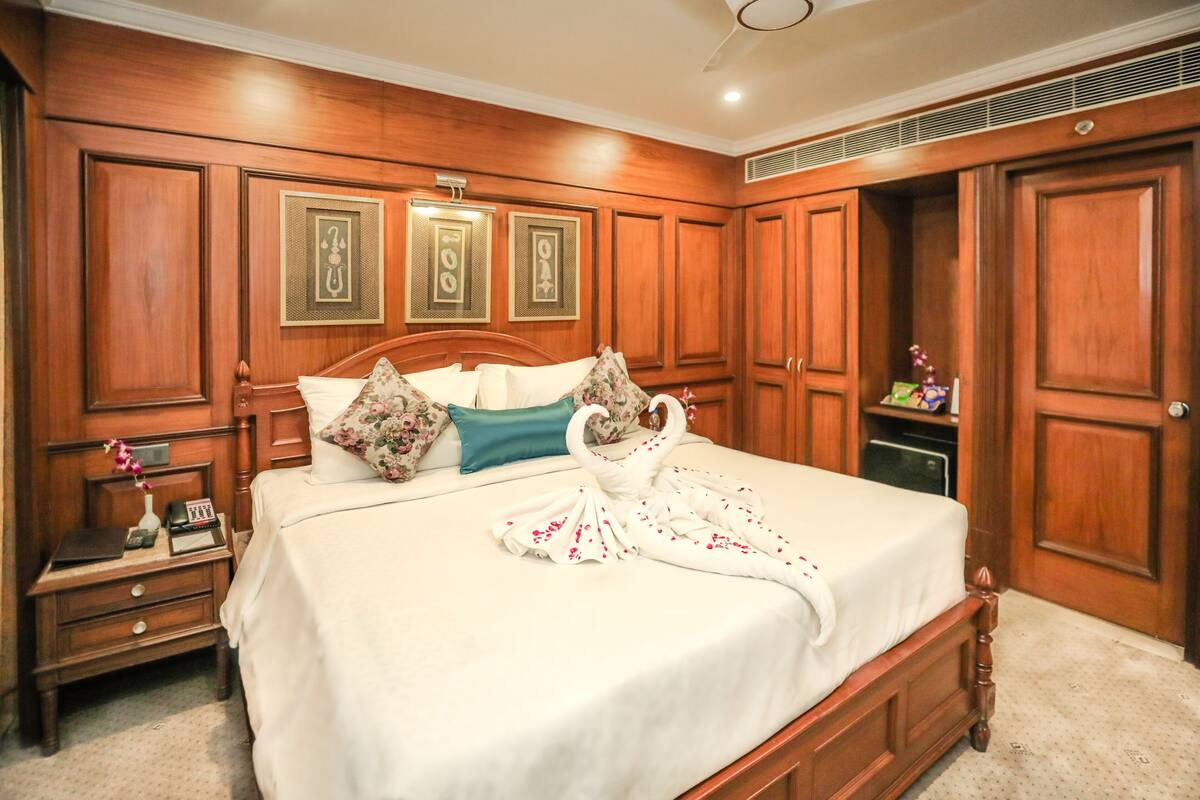 A spacious bedroom with a large bed, wooden paneling, and a nightstand with a vase and clock.