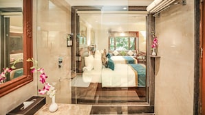 A modern hotel room with a glass partition, a bed with pillows and a blanket, a bedside table with a vase of flowers, and a large mirror. - Regenta Central Lucknow by Royal Orchid Hotels Limited (Lucknow)